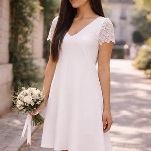 May include: A white V-neck dress with lace sleeves. The dress is knee-length and has a simple, elegant design. A bouquet of white roses and greenery is held in the hand. The background is a blurred outdoor scene.