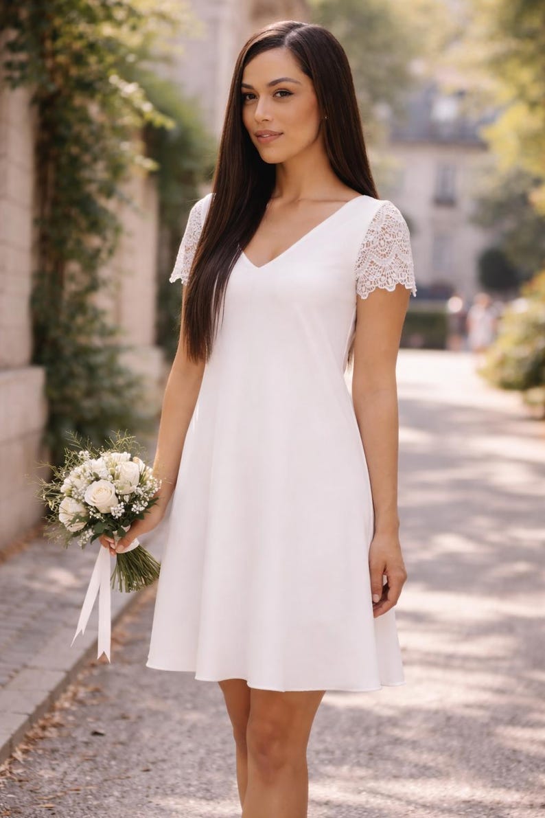 May include: A white V-neck dress with lace sleeves. The dress is knee-length and has a simple, elegant design. A bouquet of white roses and greenery is held in the hand. The background is a blurred outdoor scene.