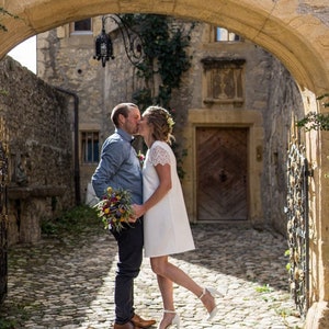 May include: A couple shares a kiss under a stone archway. The woman wears a white dress and heels, while the man is in a blue shirt and dark pants. They hold a bouquet of colorful flowers. The scene is set on a cobblestone street.