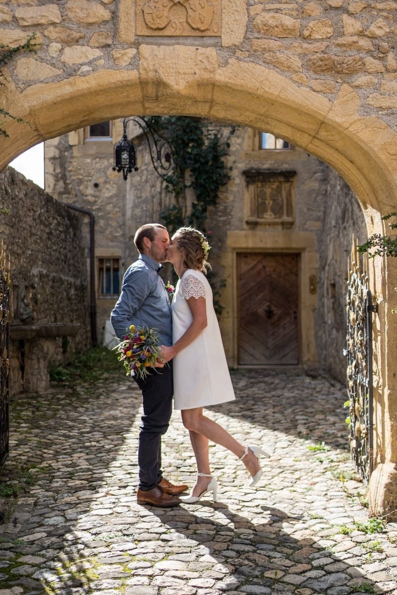 May include: A couple shares a kiss under a stone archway. The woman wears a white dress and heels, while the man is in a blue shirt and dark pants. They hold a bouquet of colorful flowers. The scene is set on a cobblestone street.