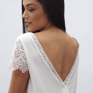 May include: A white top with a deep V-neck back and lace detailing. The short sleeves are made of lace. The top is a solid white color.