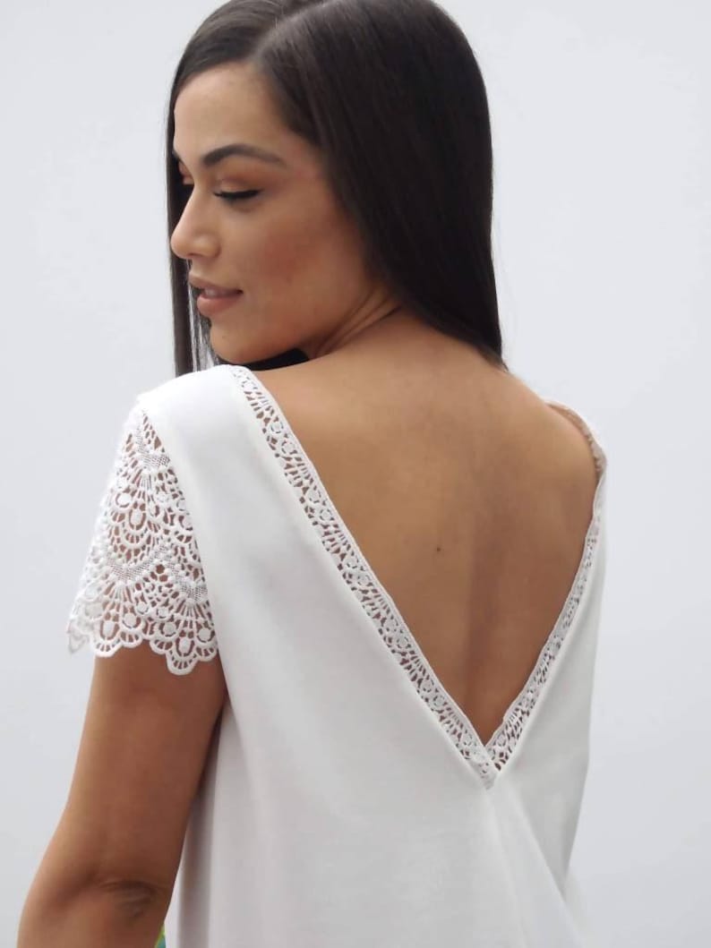 May include: A white top with a deep V-neck back and lace detailing. The short sleeves are made of lace. The top is a solid white color.