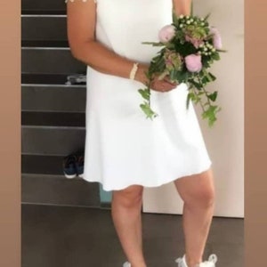 May include: A white V-neck dress with lace sleeves. The dress is paired with white sneakers with bows. A bouquet of pink and green flowers is held in front of the dress. The background includes a staircase.