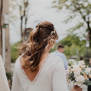 May include: A bride in a white dress with a lace-trimmed neckline and long sleeves, holding a bouquet of light-colored flowers. Her hair is styled with a braid and adorned with floral accents. A man in a white shirt walks beside her.
