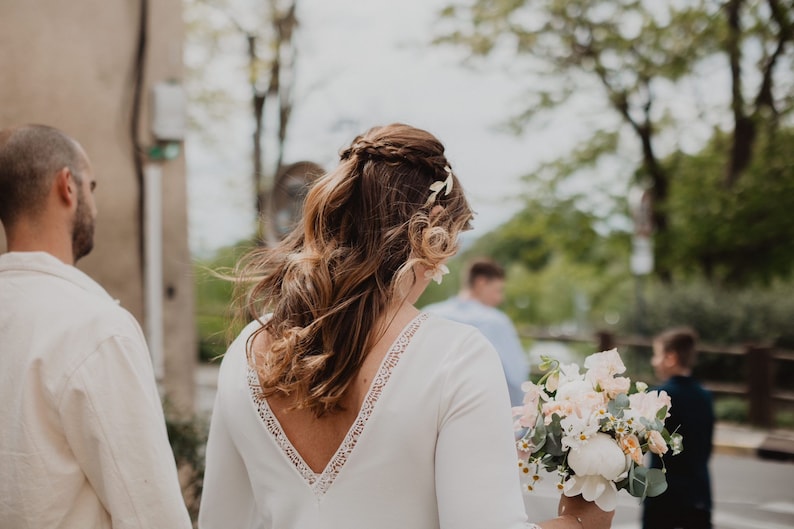 May include: A bride in a white dress with a lace-trimmed neckline and long sleeves, holding a bouquet of light-colored flowers. Her hair is styled with a braid and adorned with floral accents. A man in a white shirt walks beside her.