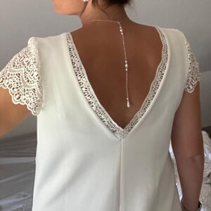 May include: A white wedding dress with a V-neckline and lace detailing on the sleeves and neckline. A delicate pearl necklace hangs down the back. The dress is a simple, elegant design.
