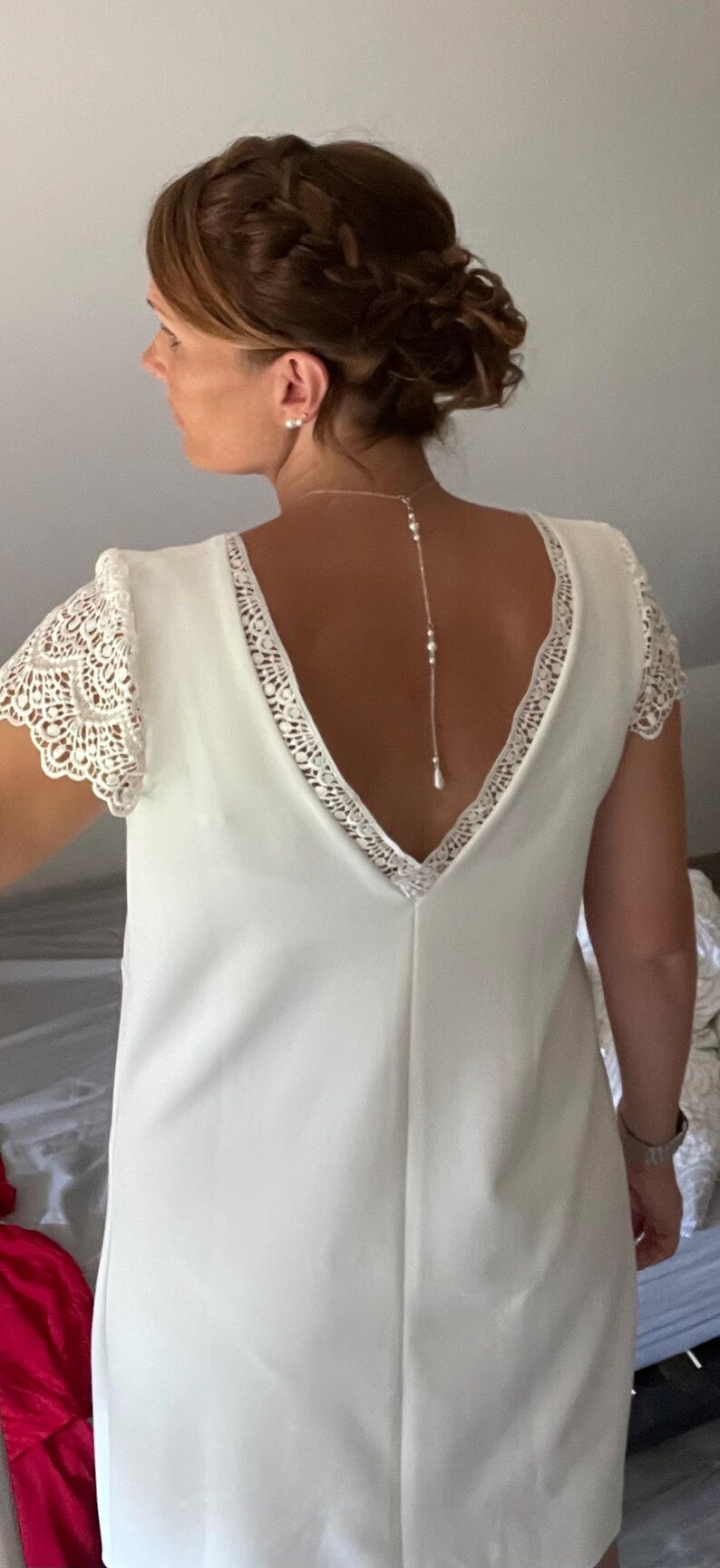 May include: A white wedding dress with a V-neckline and lace detailing on the sleeves and neckline. A delicate pearl necklace hangs down the back. The dress is a simple, elegant design.