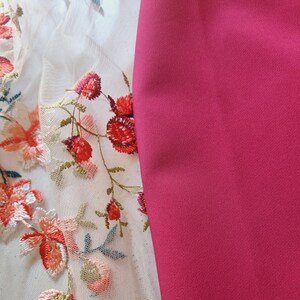 Pu&ograve; includere: Primo piano di un tessuto bianco con ricamo floreale rosso e rosa. Il tessuto &egrave; accanto a un tessuto rosa solido.