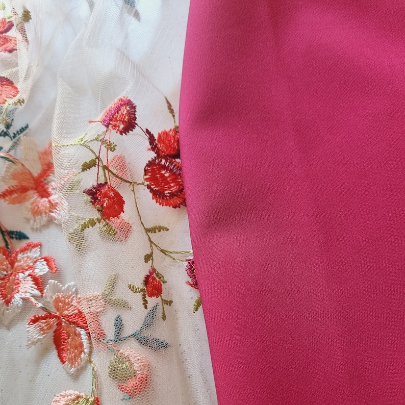 Pu&ograve; includere: Primo piano di un tessuto bianco con ricamo floreale rosso e rosa. Il tessuto &egrave; accanto a un tessuto rosa solido.