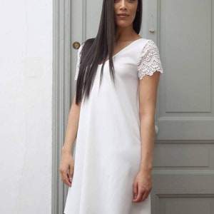 May include: A white V-neck dress with lace sleeves. The dress is knee-length and has a simple, elegant design. The model is standing in front of a gray door.