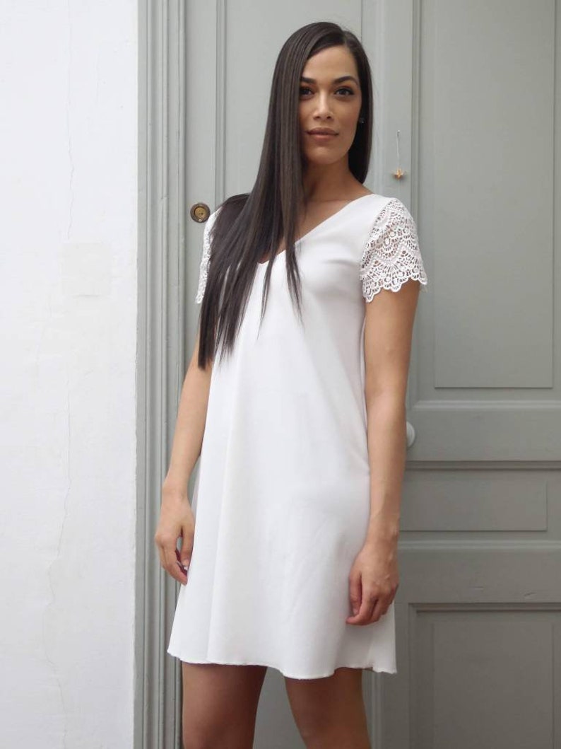 May include: A white V-neck dress with lace sleeves. The dress is knee-length and has a simple, elegant design. The model is standing in front of a gray door.