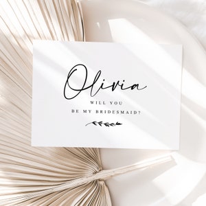 Puede incluir: Una tarjeta blanca con el nombre "Olivia" en elegante escritura negra, seguido de "Will you be my bridesmaid?" en una fuente sencilla. Una pequeña ilustración botánica en la parte inferior. La tarjeta está sobre un fondo neutro y texturizado.