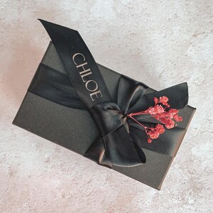 May include: A black gift box tied with a black ribbon, featuring the name "CHLOE" in gold lettering. The box is adorned with a bow and small, dried red flowers for decoration.