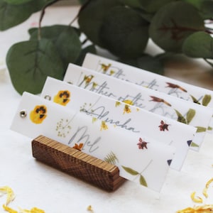 Puede incluir: Cuatro tarjetas de lugar blancas con flores prensadas y nombres escritos a mano, incluyendo "Mel" y "Madre de la novia", se muestran en un soporte de madera.