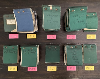 May include: A collection of vintage ledgers from 1913 to 1922, with green and blue covers. Each ledger is labeled with handwritten numbers and dates. The ledgers are arranged on a dark wooden surface.
