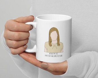 Puede incluir: Taza de cerámica blanca con un dibujo lineal de una persona con cabello rubio y una blusa color beige. El texto "WITHOUT MY CATS I AM UTTERLY AND COMPLETELY ALONE" está impreso debajo. La taza tiene un asa curva.