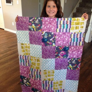 May include: A patchwork quilt featuring a range of colourful fabrics, including purple, green, yellow and white. The quilt is made up of squares with different patterns and designs.