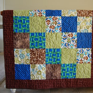 May include: A brown, green and blue patchwork quilt with a quilted pattern. The quilt is made up of 16 squares, each with a different pattern. The quilt is hanging on a wall.
