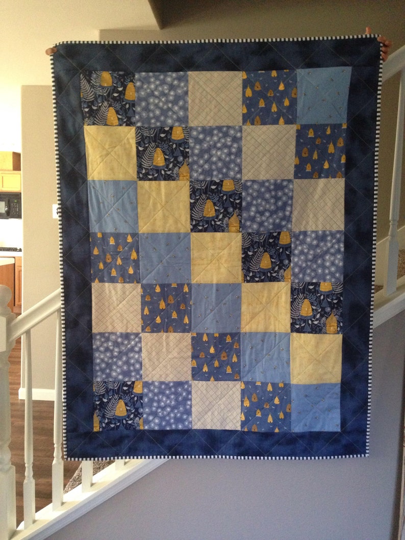 May include: A blue and yellow patchwork quilt with a white border. The quilt is made of squares of different fabrics, each with a different pattern. The quilt is hanging on a wall.