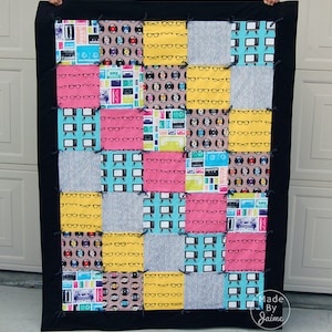 May include: A patchwork quilt with a black border. The quilt is made of various fabrics with different patterns, including eyeglasses, records, and televisions. The quilt is made in a grid pattern with 16 squares. The quilt is labelled "Made By Jaime".
