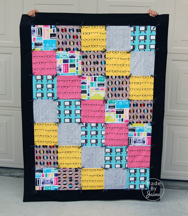 May include: A patchwork quilt with a black border. The quilt is made of various fabrics with different patterns, including eyeglasses, records, and televisions. The quilt is made in a grid pattern with 16 squares. The quilt is labelled "Made By Jaime".