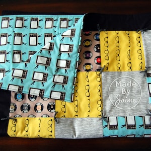 May include: A patchwork quilt featuring a range of fabrics, including a blue fabric with a pattern of televisions, a yellow fabric with a pattern of spectacles, a grey fabric with the text "Made By Jaime", and a pink fabric with a pattern of spectacles.