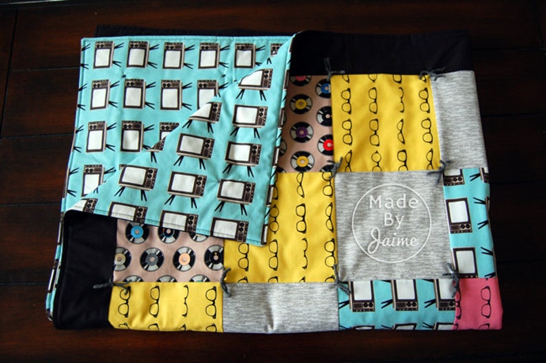 May include: A patchwork quilt featuring a range of fabrics, including a blue fabric with a pattern of televisions, a yellow fabric with a pattern of spectacles, a grey fabric with the text "Made By Jaime", and a pink fabric with a pattern of spectacles.