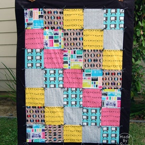May include: A patchwork quilt with a black border. The quilt is made of various fabrics with different patterns, including eyeglasses, records, and televisions. The quilt is made in a grid pattern with 16 squares. The quilt is labelled "Made by Jaime".