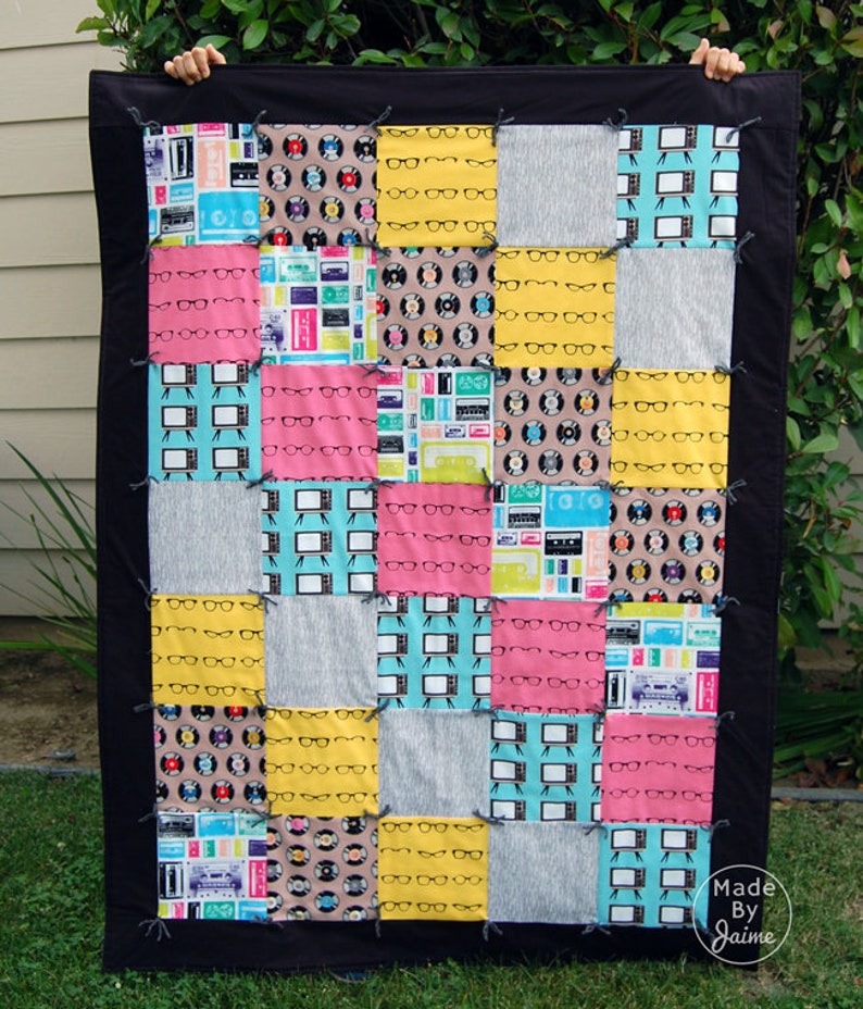 May include: A patchwork quilt with a black border. The quilt is made of various fabrics with different patterns, including eyeglasses, records, and televisions. The quilt is made in a grid pattern with 16 squares. The quilt is labelled "Made by Jaime".