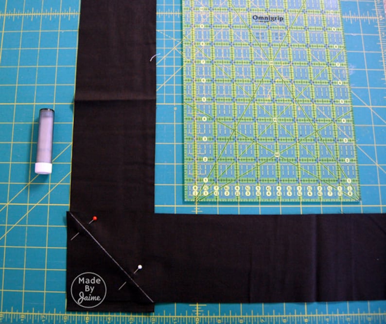 May include: A close-up of a black fabric corner being sewn with pins and a ruler. The fabric is on a green cutting mat. The text "Made By Jaime" is visible in the bottom left corner.
