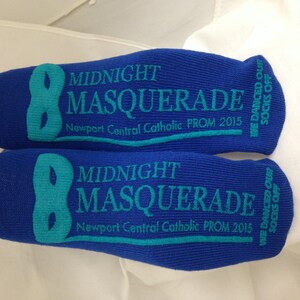 May include: A pair of blue socks with the text "Midnight Masquerade" and "Newport Central Catholic PROM 2015" printed on them. The socks also have a teal mask design and the text "We danced our socks off".