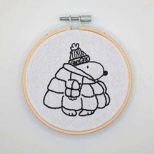 May include: Embroidered hoop art featuring a black and white illustration of a small animal wearing a hat and a coat. The animal is standing on a white background.
