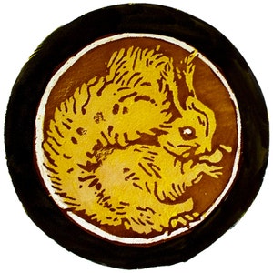 Könnte beinhalten: Eine runde, schwarze und braune Keramikfliese mit weißem Rand. Die Fliese zeigt eine gelbe Silhouette eines Eichhörnchens mit buschigem Schwanz.