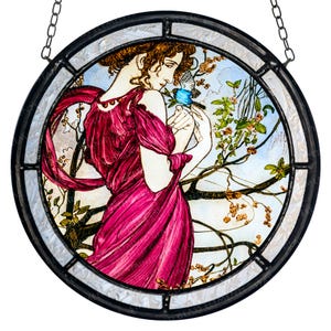 May include: Round stained glass panel featuring a woman in a flowing magenta dress, holding a blue bird. The artwork is set against a backdrop of a light blue sky and flowering branches, framed by a black metal border and hanging chain.