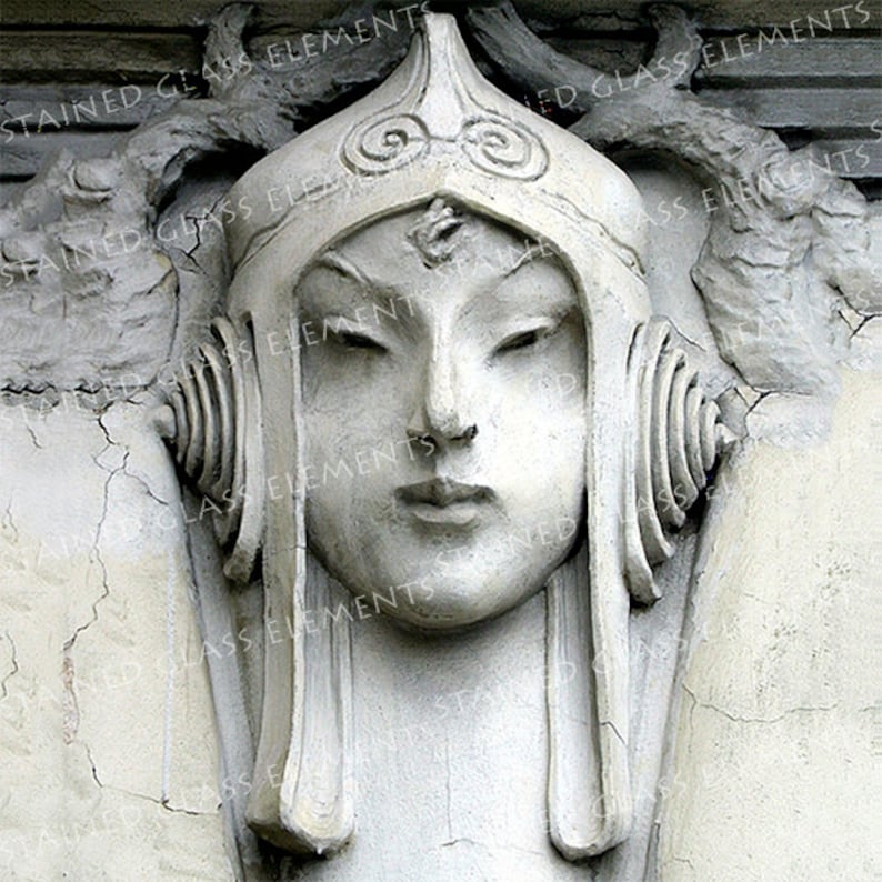 May include: A white plaster sculpture of a woman's face with a crown and elaborate headdress. The sculpture is mounted on a wall.