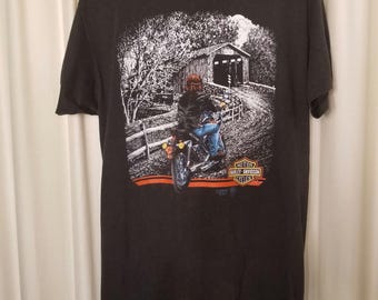 May include: A black short-sleeve t-shirt featuring a graphic print of a person riding a motorcycle toward a covered bridge. The design includes the Harley-Davidson Motor Cycles logo in orange and white, with trees and a fence in the background.