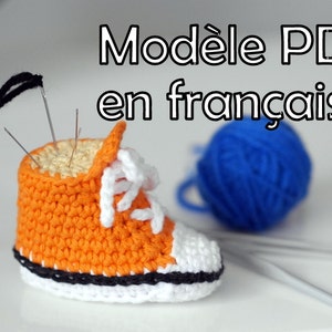 May include: An orange and white crocheted sneaker-shaped pincushion with needles inserted. The text "Modèle PDF en français" is visible. A blue ball of yarn and crochet hooks are in the background. The sneaker has white laces and a black sole.