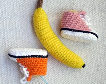 Puede incluir: Un plátano amarillo tejido a ganchillo con un tallo marrón y dos zapatillas tejidas a ganchillo, una rosa y otra naranja, con suelas blancas y ribetes negros. Las zapatillas son en miniatura y parecen hechas a mano.