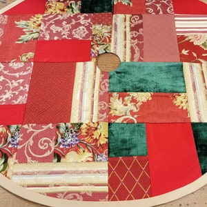 May include: A round patchwork Christmas tree skirt with a beige border. The skirt is made of various fabrics in shades of red, green, and gold with floral and geometric patterns.