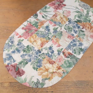 May include: Oval shaped table runner with a floral pattern. The pattern features pink, yellow, blue, and green flowers on a white background.