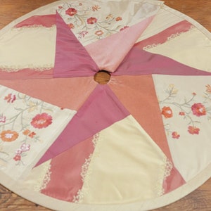 May include: A circular, cream-colored tree skirt with a pink and peach patchwork design. The skirt has a scalloped edge and is decorated with embroidered floral patterns.
