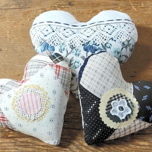 May include: Three heart-shaped decorative pillows. The top pillow is white with a floral pattern and lace trim. The other two pillows are patchwork, featuring various fabric patterns and colors, with decorative appliqués.