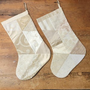 May include: Two beige Christmas stockings with patchwork designs. Each stocking features a variety of fabrics in cream, tan, and ivory, with different textures and patterns. They have a loop for hanging and are set against a wooden surface.