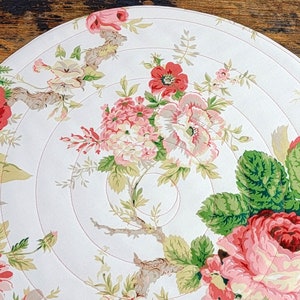 May include: A round fabric placemat featuring a white background with a floral pattern of red, pink, and white roses, green leaves, and brown branches. The placemat is constructed with circular quilted stitching that follows the round shape.