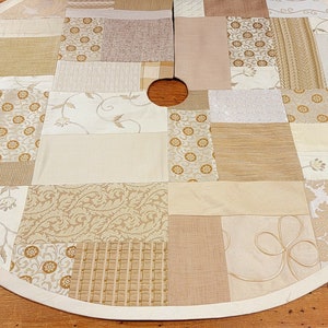 May include: A cream-colored patchwork tree skirt with a variety of beige and brown patterns. The skirt has a white trim around the edge and a hole in the center for the tree trunk.