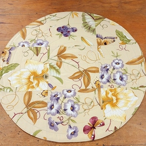 May include: A round table runner with a floral design. The runner is made of fabric and features a variety of flowers, leaves, and butterflies in shades of yellow, green, and purple. The background is a cream color.