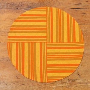 May include: A round placemat with a striped pattern in shades of orange, yellow, and brown. The design is divided into four quadrants, with alternating vertical and horizontal stripes. The placemat is placed on a wooden surface.