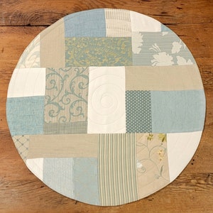 May include: A round, patchwork table runner with a variety of blue and beige fabrics. The runner is made of different patterns, including floral, geometric, and striped designs.