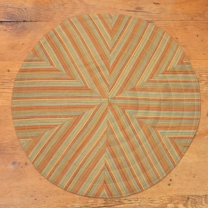 May include: A round, multi-colored placemat with a geometric pattern of stripes and triangles. The placemat is made of a woven fabric and has a beige, brown, and green color scheme.