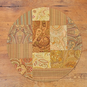 May include: A round, multi-colored patchwork table runner with a paisley pattern. The runner is made of fabric and has a variety of brown, beige, and gold tones.